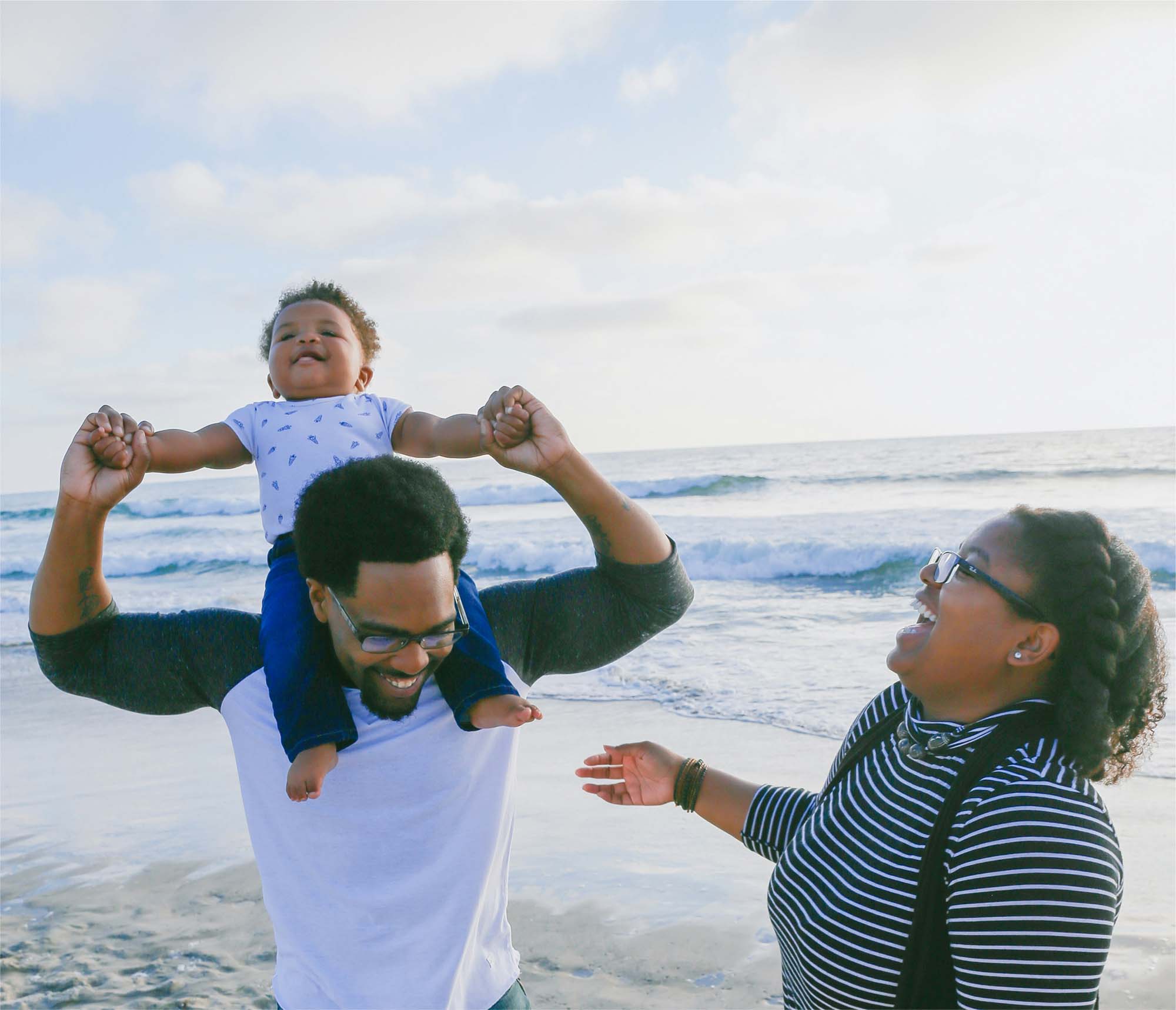 A father carrying his baby on his shoulders at the beach while the baby's mother laughs