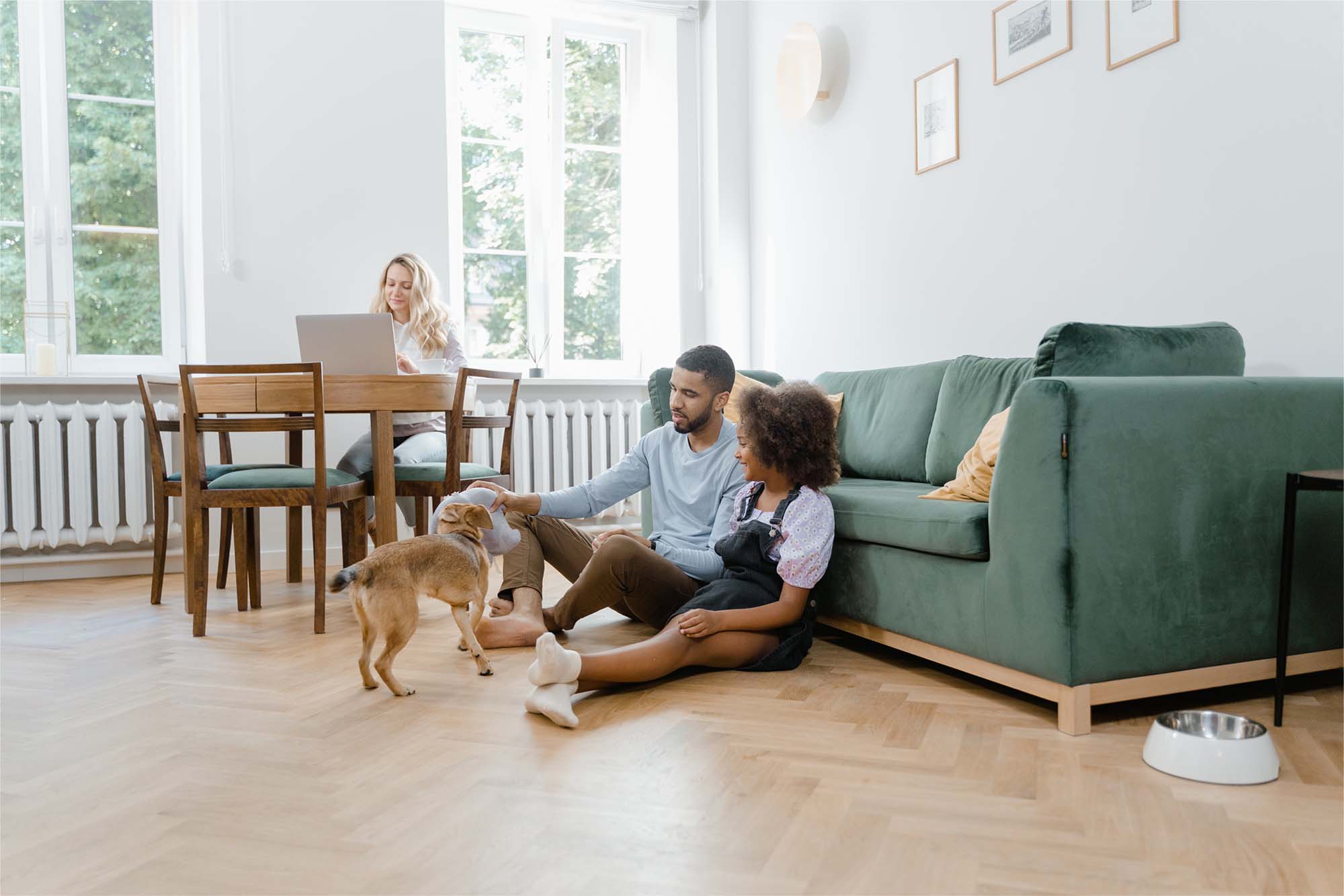 A family at home: a father and his daughter play with their dog while the mother works at the dining table