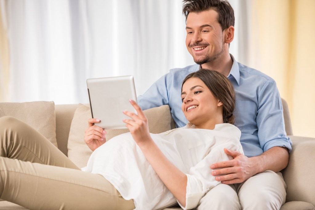 An expecting couple relaxing on a sofa while they look at a tablet together